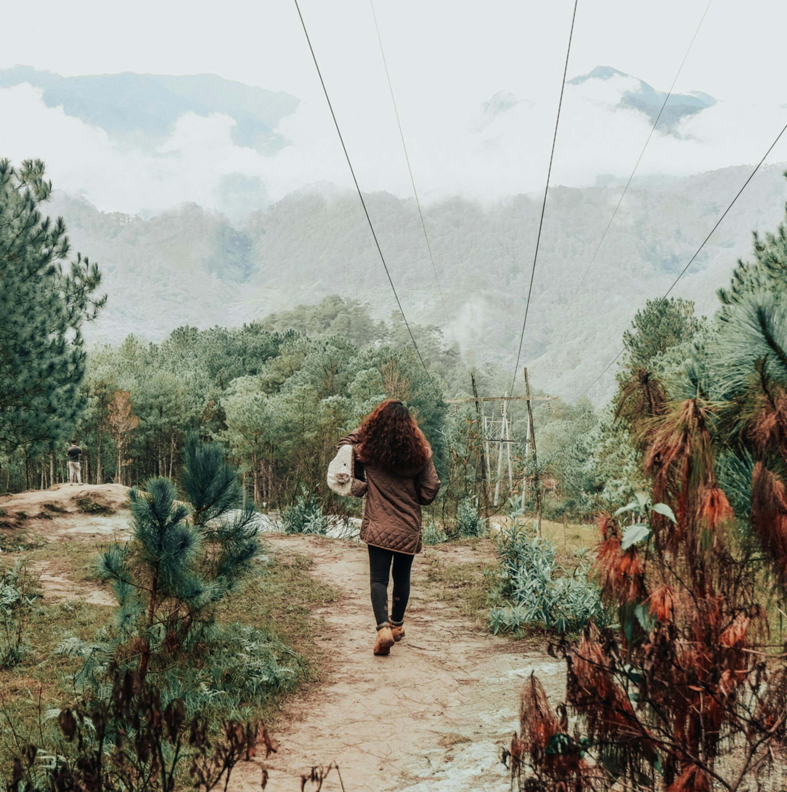 A person walking on a dirt path in Sagada, surrounded by lush greenery and distant mountains.
