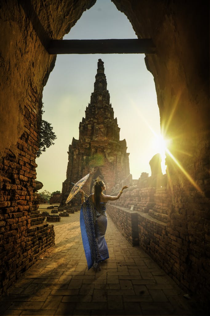 A woman in traditional wear stands with an umbrella at a historic Ayutthaya temple during sunset.