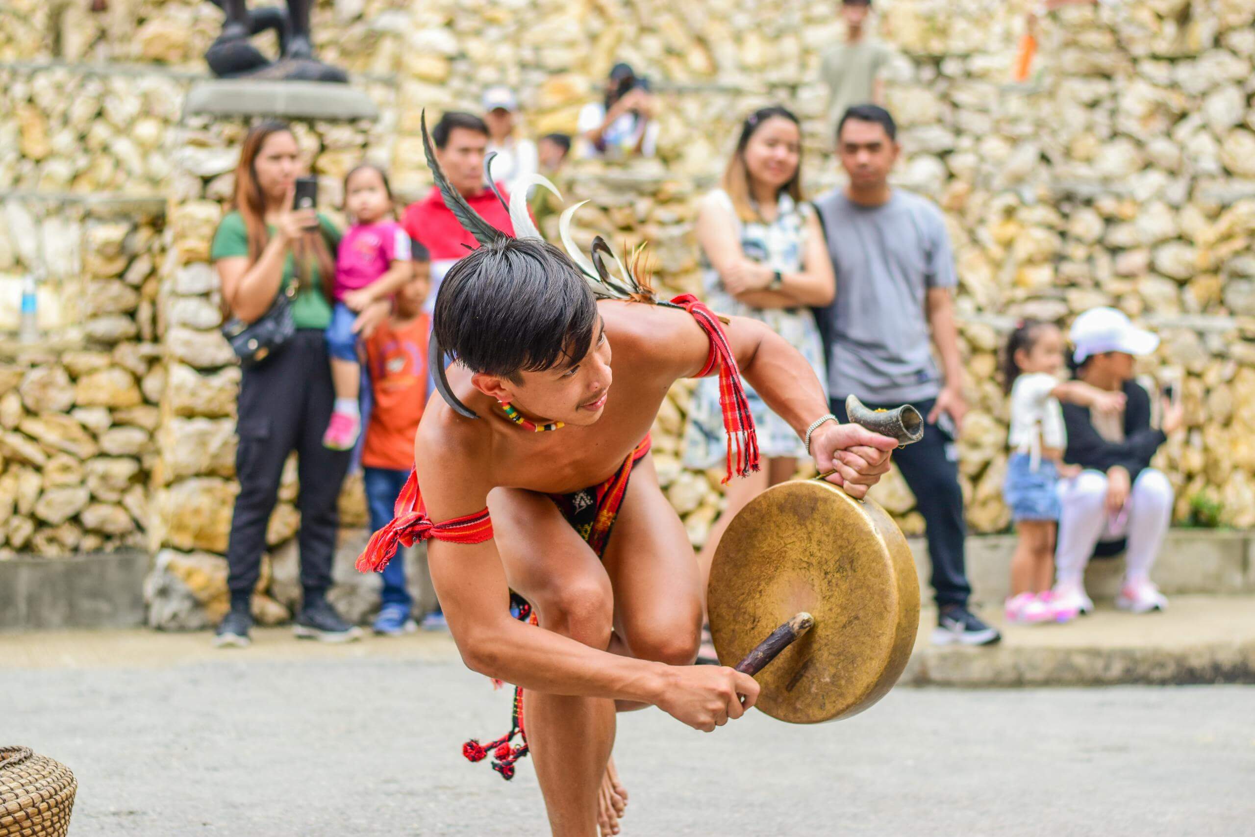 A teenager in traditional attire energetically plays a drum during a festival, engaging an audience.