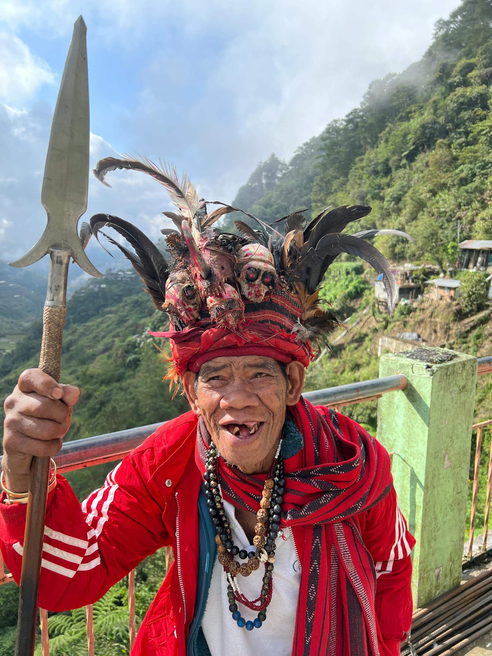 Elderly man in traditional Ifugao costume holding a spear against a scenic Banaue backdrop.