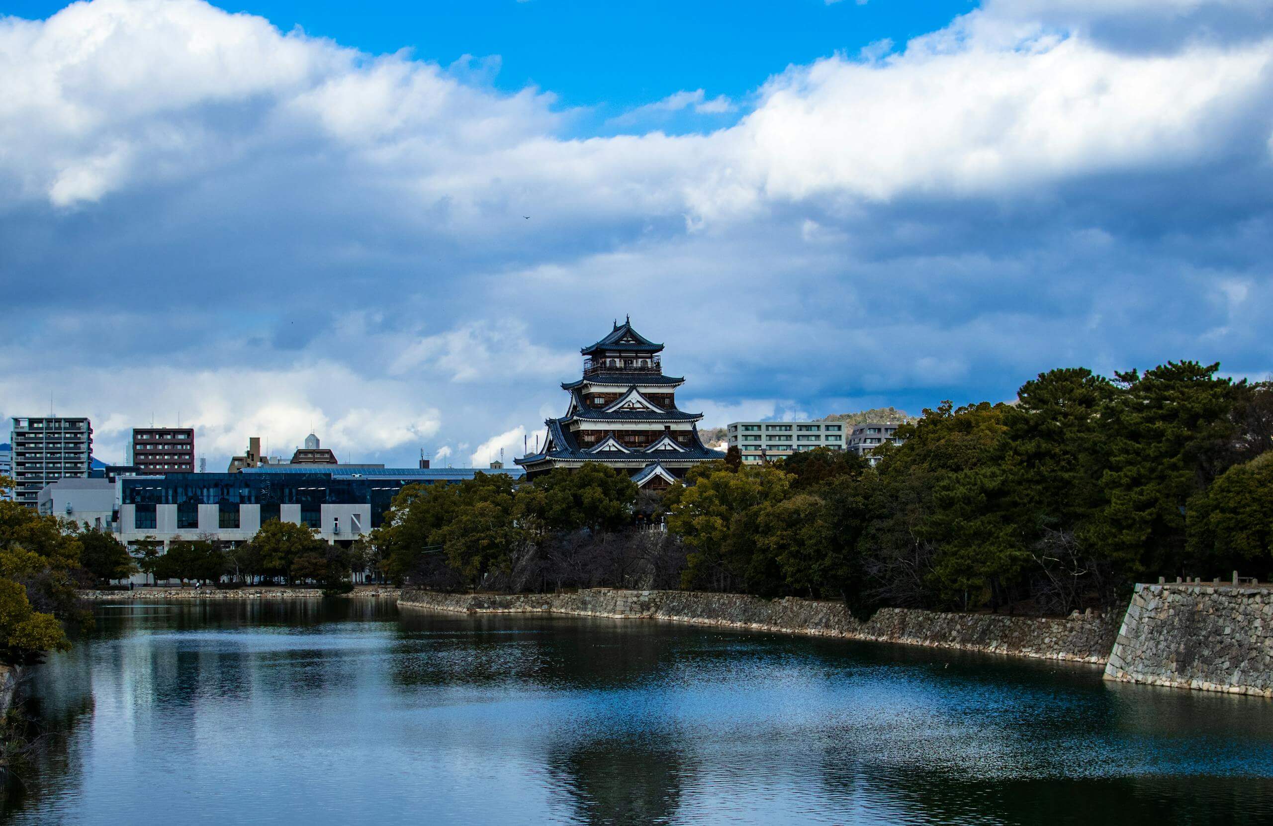 Peaceful reflection of Hiroshima Castle surrounded by trees and water under a dramatic sky.