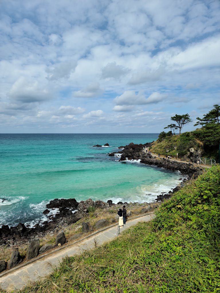A picturesque view of a turquoise sea and rocky coastline in Jeju, South Korea, under a partly cloudy sky.