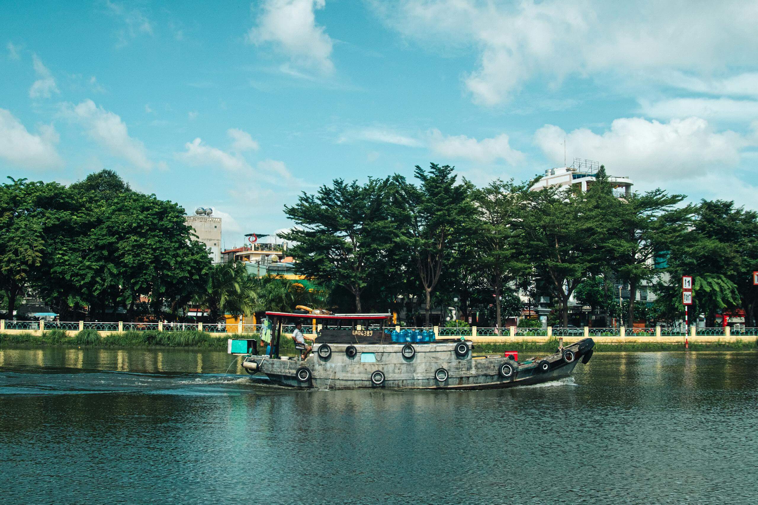 A riverboat sails on the Saigon River in Ho Chi Minh City, surrounded by lush greenery and urban buildings.