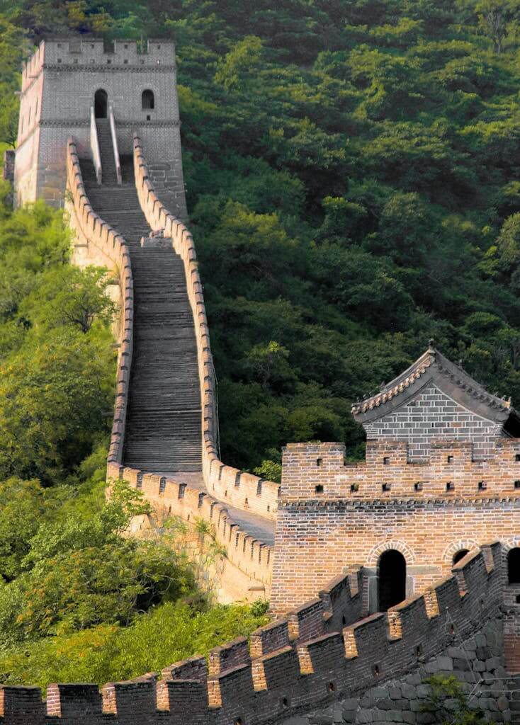 A scenic view of the Great Wall of China curving over lush, green hills in bright daylight.