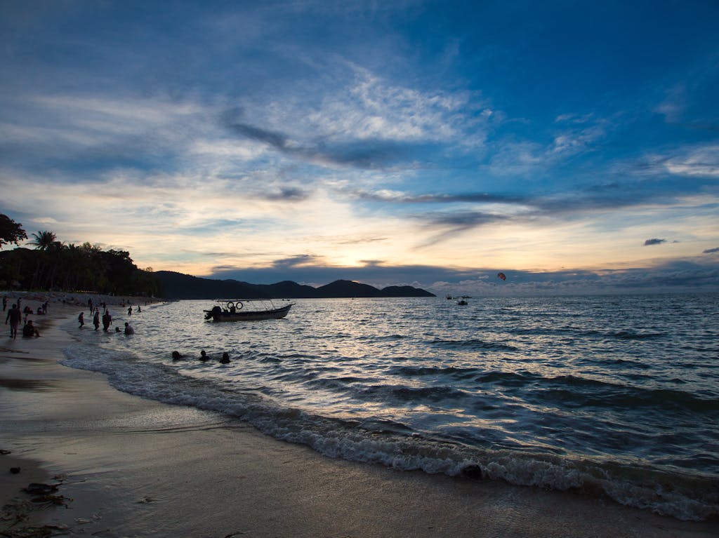 A serene sunset view over Batu Ferringhi Beach in Penang, Malaysia, with silhouettes and waves.