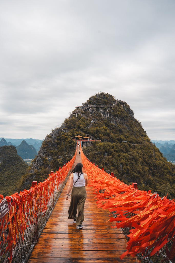 A woman crosses a vibrant red ribbon bridge toward a lush mountain in Guilin, China, under a cloudy sky.