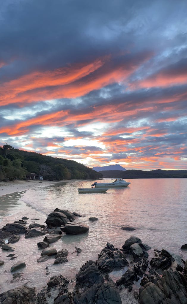 Breathtaking sunset view on a rocky beach in Kudat, Sabah, Malaysia with boats and vibrant skies.