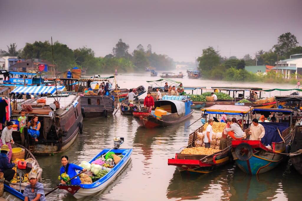 Lively floating market scene with locals trading fresh produce from traditional boats on a calm river. Mekong Delta Vietnam Tour