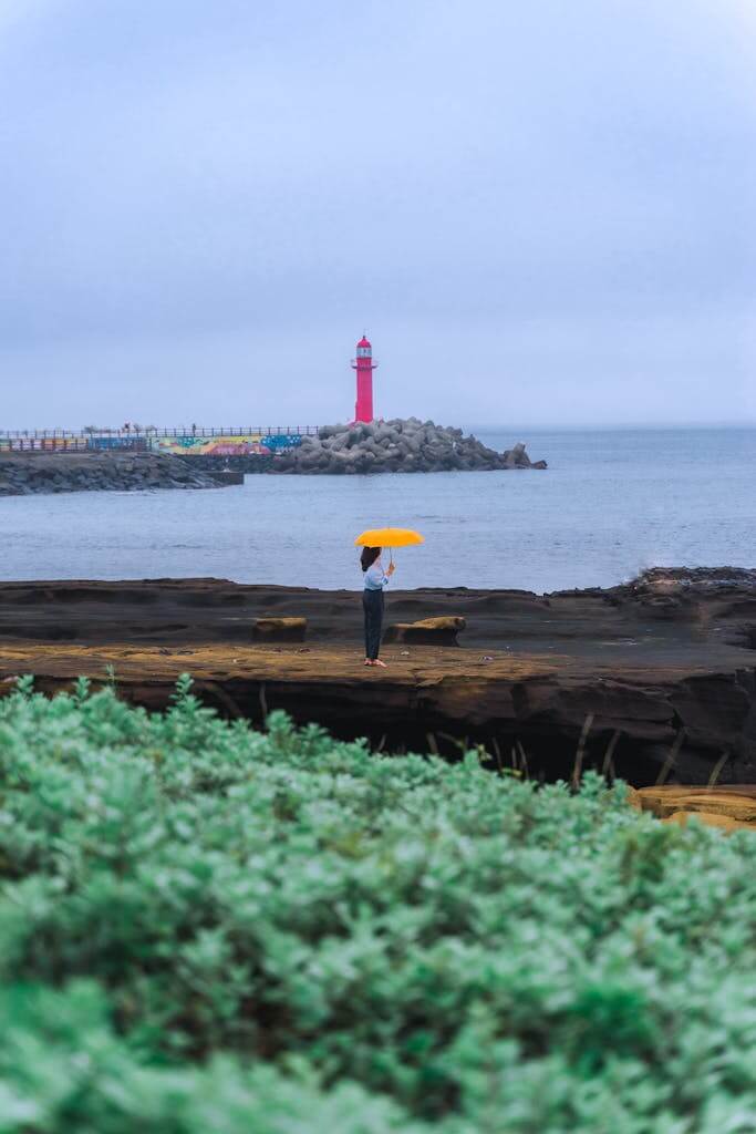 Person standing with a yellow umbrella by a red lighthouse on Jeju Island's rocky coast.