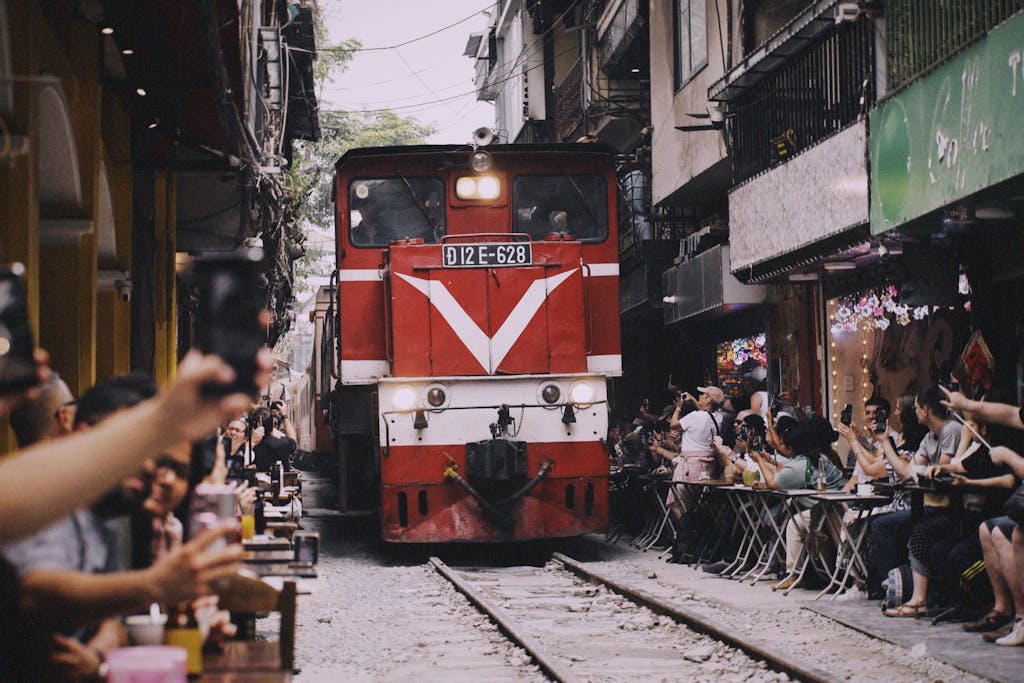 Red train moves through narrow Hanoi street as people capture the moment with their phones.