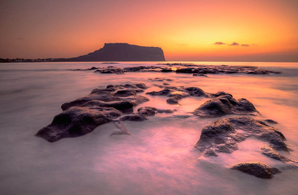 Stunning sunrise over Seongsan Ilchulbong Peak, Jeju Island, South Korea, featuring smooth ocean and rocky foreground.
