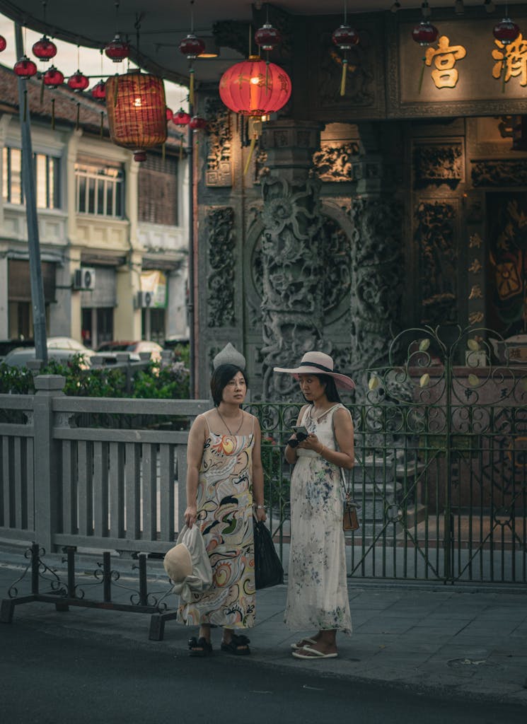 Two women in elegant dresses stand near a historical temple in George Town, Penang.